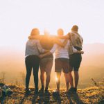 Culture Community - four person hands wrap around shoulders while looking at sunset