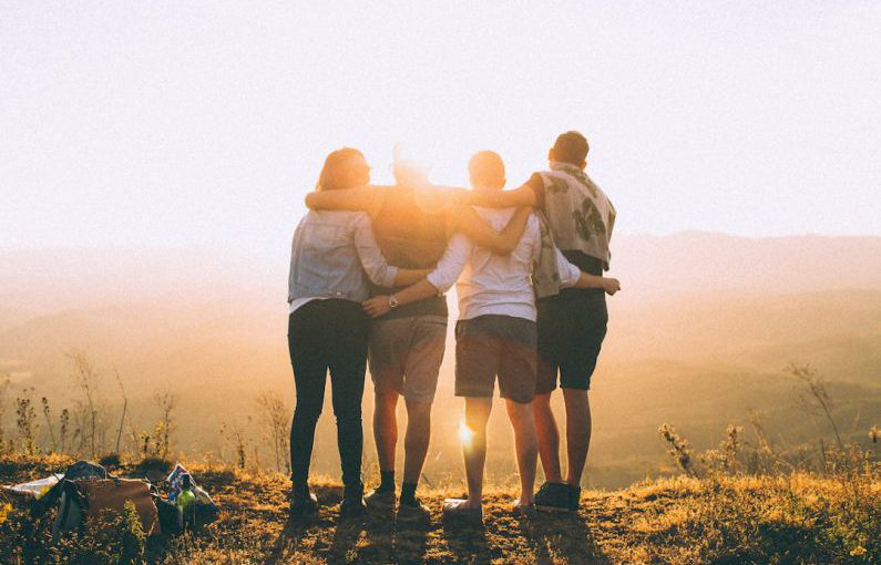 Culture Community - four person hands wrap around shoulders while looking at sunset